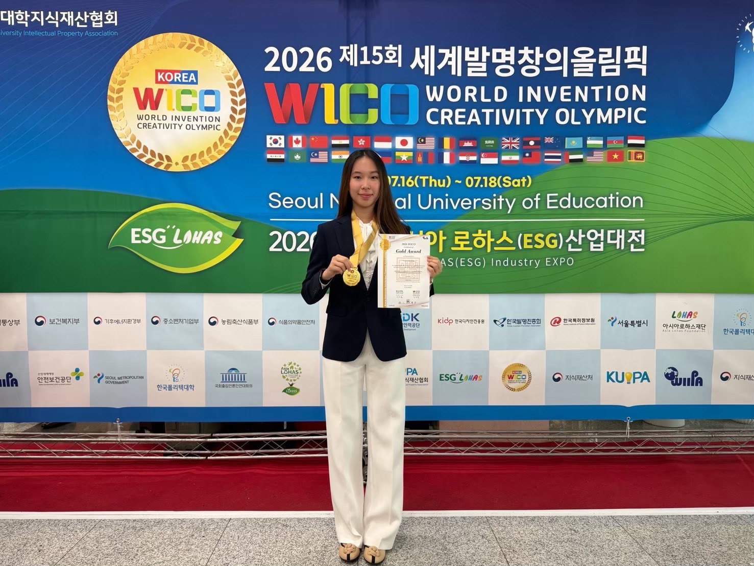 Winnie holding her WICO 2026 Gold Award certificate and medal in front of the event banner